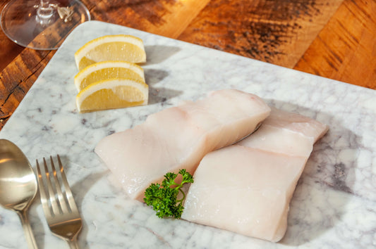 Two fresh halibut fillets on a marble board with lemon wedges, parsley and cutlery